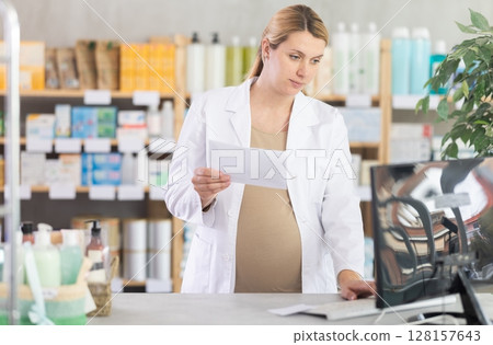 Pregnant woman employee in white coat works in pharmacy sales area using computer. Pregnant woman employee in white coat works in pharmacy sales area using computer. 128157643