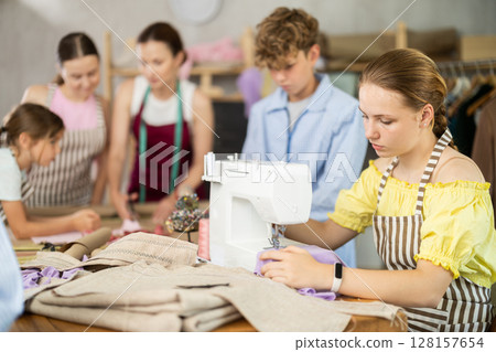 Teenage girl works on sewing machine while teacher and children learn how to cut fabric 128157654