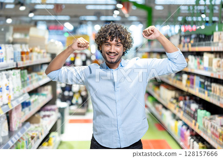 A happy man with curly hair in a supermarket raises his arms in celebration, surrounded by shelves. A happy man with curly hair in a supermarket raises his arms in celebration, surrounded by shelves. 128157666