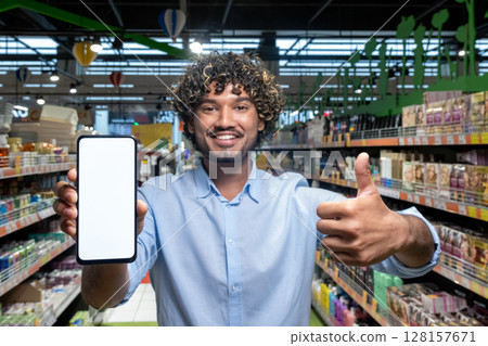 A man holding a phone with a blank screen in a supermarket, giving a thumbs-up, promoting online shopping. 128157671