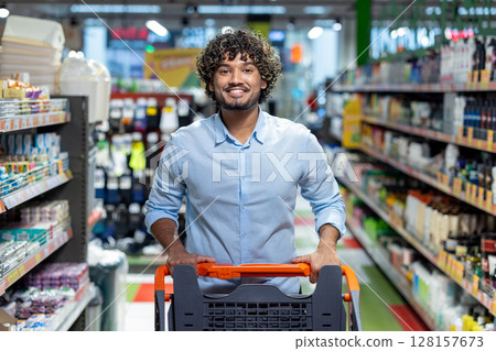 A smiling man pushes a shopping cart down an aisle in a brightly lit grocery store, focused on purchasing. 128157673
