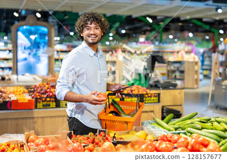 A smiling man shops for fresh produce in a well-lit grocery store, holding a tomato and shopping basket. A smiling man shops for fresh produce in a well-lit grocery store, holding a tomato and shopping basket. 128157677