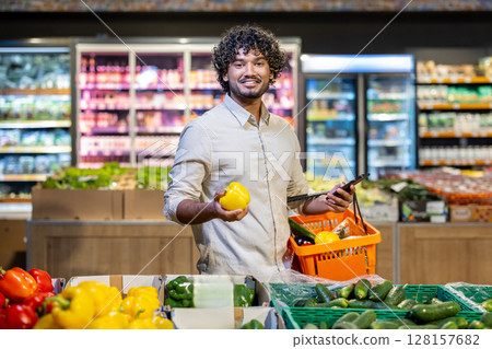 A smiling man holding a yellow bell pepper while shopping in a supermarket. He is looking at the camera, carrying a basket of groceries. A smiling man holding a yellow bell pepper while shopping in a supermarket. He is looking at the camera, carrying a basket of groceries. 128157682