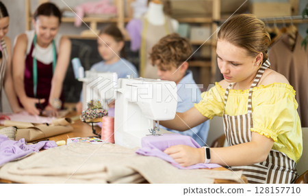 Teenage girl works on sewing machine while teacher and children learn how to cut fabric 128157710
