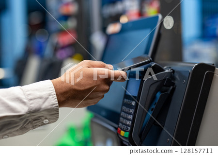 A person uses their smartphone to make a contactless payment at a point-of-sale terminal in a retail setting. The focus is on the hand and the device. 128157711