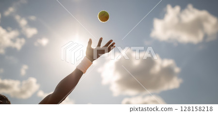 Tennis player tossing ball in bright sunlight with cloudy sky background, capturing action moment of serve preparation with wristband on arm 128158218