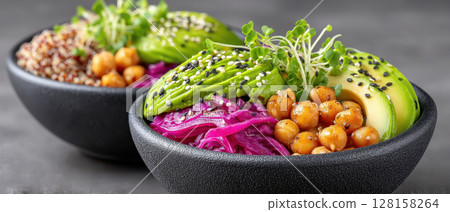 Vegan Buddha bowl with chickpeas, avocado slices, quinoa, purple cabbage, and microgreens topped with black and white sesame seeds, served in black bowls on gray surface 128158264