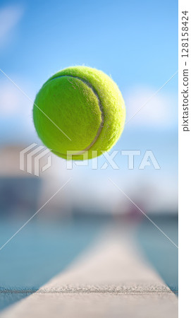 Tennis ball bouncing on hard court with dynamic motion and bright blue sky background creating energetic and vibrant atmosphere 128158424