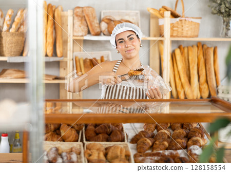 Young female seller putting croissant in bag Young female seller putting croissant in bag 128158554