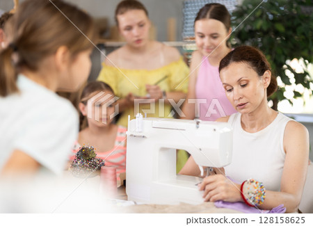 Woman teacher sews product surrounded by students Woman teacher sews product surrounded by students 128158625