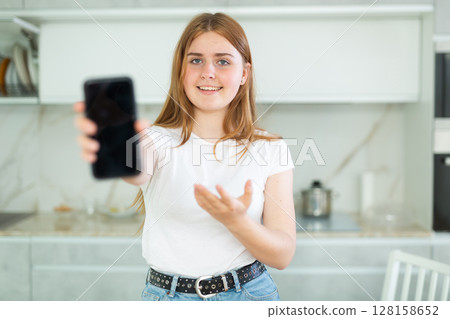 Smiling teenage girl presenting smartphone with blank black screen with copy space in modern cuisine. Free copy space for your design or advertisement Smiling teenage girl presenting smartphone with blank black screen with copy space in modern cuisine. Free copy space for your design or advertisement 128158652