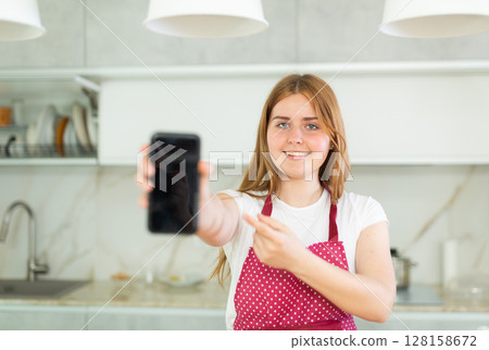 Happy teen girl in red polka dot apron showing modern smartphone with black screen in modern kitchen 128158672