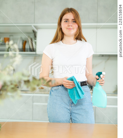 in kitchen, woman hostess in casual clothes stands and holds rag and cleaning spray in hands. in kitchen, woman hostess in casual clothes stands and holds rag and cleaning spray in hands. 128158694