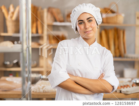 Young female baker stands in bake house, ready to greet customers Young female baker stands in bake house, ready to greet customers 128158697