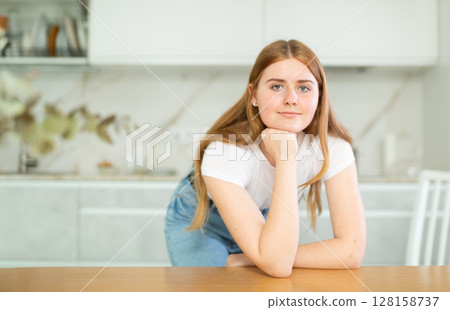 Portrait of positive teenage girl smiling at camera while leaning on countertop in cozy kitchen 128158737