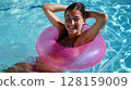 Cheerful young woman in a pink rubber swimming pool ring, relaxing at a hotel club swimming pool on a sunny summer day, capturing the joy and fun of her vacation in the clear blue water 128159009