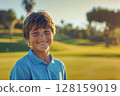 Young caucasian boy at a golfing training lesson, beaming with joy as he looks at the camera, standing on a beautiful golf course, embracing the excitement of learning to play golf amidst a 128159019