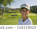 Young caucasian boy at a golfing training lesson, beaming with joy as he looks at the camera, standing on a beautiful golf course, embracing the excitement of learning to play golf amidst a 128159020