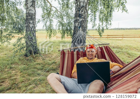man relaxing in hammock working on laptop in nature in red cap and yellow shirt with green grass background, remote work travel blog digital marketing outdoor lifestyle, copy space man relaxing in hammock working on laptop in nature in red cap and yellow shirt with green grass background, remote work travel blog digital marketing outdoor lifestyle, copy space 128159072