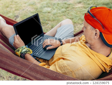 man relaxing in hammock working on laptop in nature wearing red cap and yellow shirt with green grass background concept of remote work travel blog digital marketing outdoor lifestyle 128159075