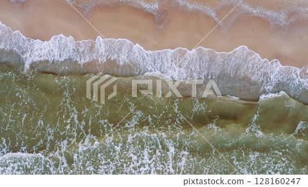 Aerial view of waves on a sandy tropical beach. Oncoming waves crashing against the sandy tropical shore over and over again. Aerial drone top view of sandy and wavy beach. 128160247