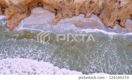 Aerial view of stormy waves beating against a rocky shore on a sandy tropical beach. Oncoming waves crashing against the sandy tropical shore over and over again. 128160259