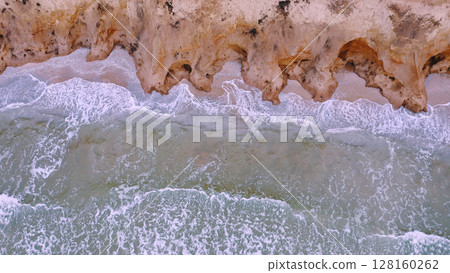 Aerial view of waves beating against a rocky shore on a sandy tropical beach. Oncoming waves crashing against the sandy tropical shore over and over again. 128160262