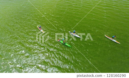 Top down view of four paddleboarders racing on vibrant green water during a stand-up paddleboarding competition with numbered jerseys and paddles. 128160290