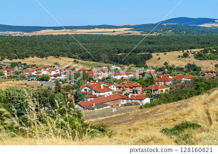 Scenic view of picturesque Bulgarian village in green hills, charming houses with red roofs, expansive fields under clear blue sky. Serene and tranquil atmosphere in summer 128160821