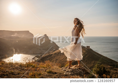 A woman stands on a hill overlooking the ocean. She is wearing a white dress and has long hair. The scene is serene and peaceful, with the sun shining brightly in the background. 128160957