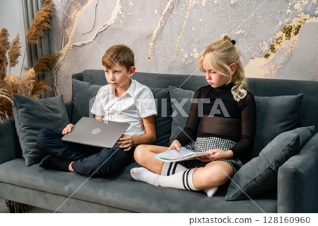 Children Laptop Couch Studying - Boy and girl on couch; boy using laptop, girl reading book. 128160960