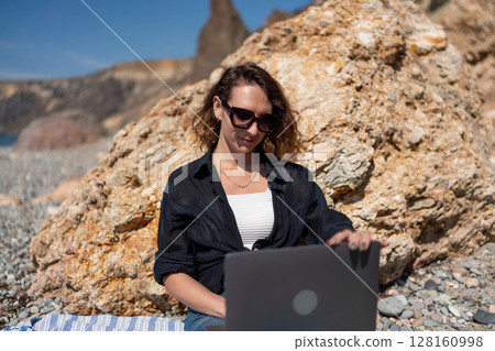 Beach laptop freelance: Woman working on laptop at sunny beach by rocks in casual clothing. 128160998