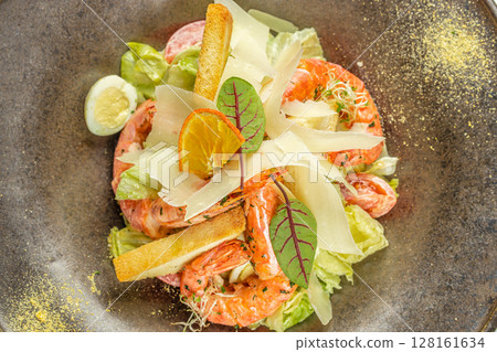 A vibrant shrimp Caesar salad served with crisp romaine lettuce, cherry tomatoes, shaved parmesan, crunchy croutons, and garnished with a slice of dried orange and fresh greens 128161634