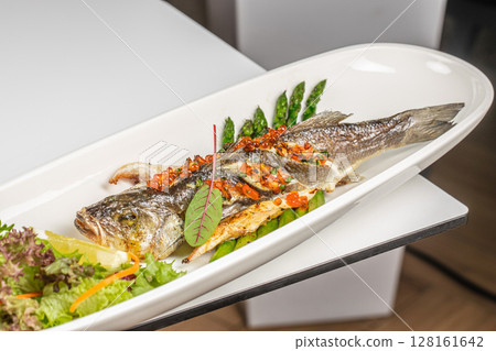 A beautifully presented whole grilled fish garnished with herbs, vegetables, red caviar, and served with fresh greens and lemon on a white plate 128161642