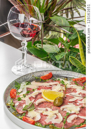 A gourmet beef carpaccio served with parmesan shavings, arugula, capers, cherry tomatoes, and lemon, paired with a glass of red wine in an elegant setting 128161651