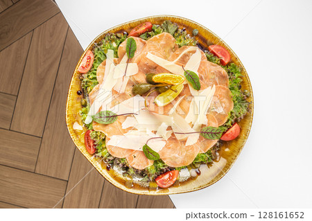 A beautifully plated salmon carpaccio with parmesan shavings, fresh greens, cherry tomatoes, capers, and lemon slices, elegantly arranged on a rustic dish A beautifully plated salmon carpaccio with parmesan shavings, fresh greens, cherry tomatoes, capers, and lemon slices, elegantly arranged on a rustic dish 128161652