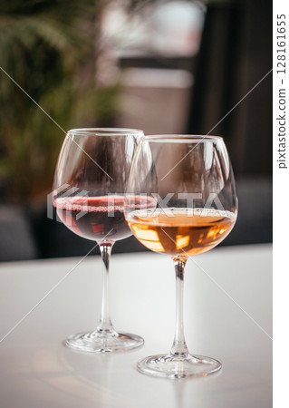 Two elegant wine glasses filled with red and amber wine stand on a white surface, reflecting soft indoor light in a cozy setting 128161655