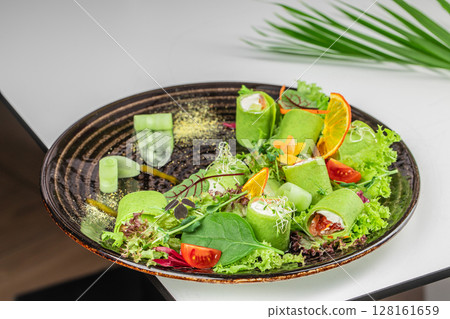 A vibrant plate of green vegetable rolls with fresh greens, cherry tomatoes, sprouts, and dried orange garnishes served on a dark ceramic dish 128161659