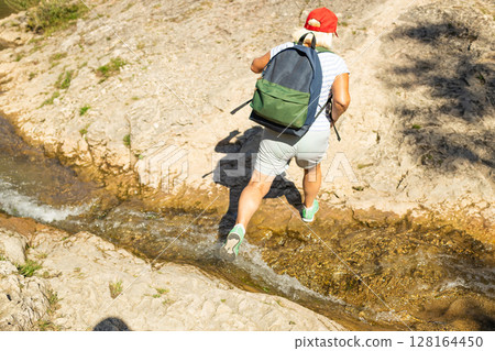 Senior Woman with Backpack Crossing Mountain Stream 128164450