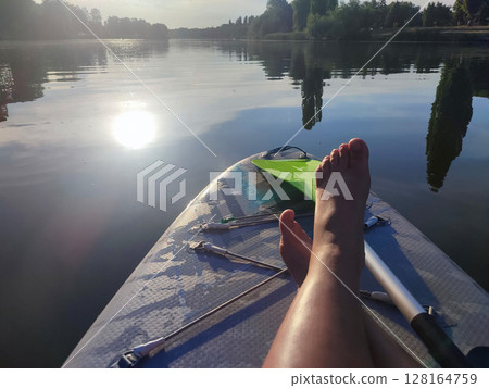 Bare legs of woman on SUP board on surface of river or lake with sun reflecting and with trees on banks on warm summer evening. Person swimming on SUP board on pond on calm day. Tourism rest sport Bare legs of woman on SUP board on surface of river or lake with sun reflecting and with trees on banks on warm summer evening. Person swimming on SUP board on pond on calm day. Tourism rest sport 128164759