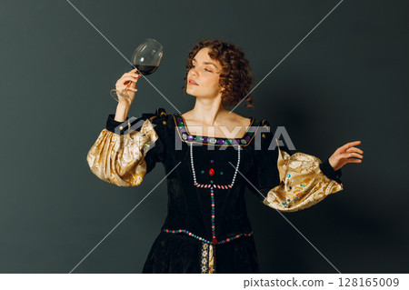 Young adult woman dressed in a medieval dress holding a glass of wine and drinking a red wine 128165009