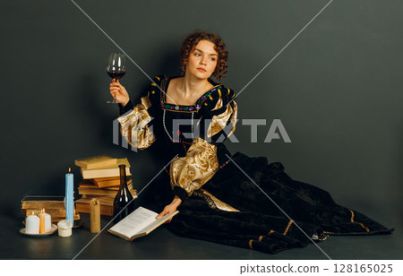 Young adult woman dressed in a medieval dress sitting near ancient books and candles with bottle of red wine and holding a glass of wine 128165025