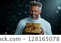 Man wearing white sweater smiling while holding a golden gift box during a snowy celebration 128165078