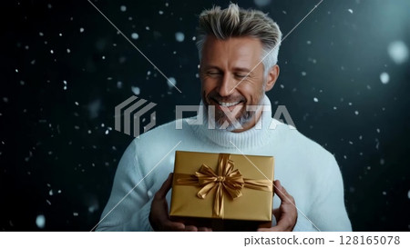 Man wearing white sweater smiling while holding a golden gift box during a snowy celebration 128165078