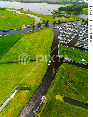 A top-down view of a marina with multiple boats docked along wooden piers, a boat cruising through a narrow waterway surrounded by green parks and natural grassy areas. 128165718