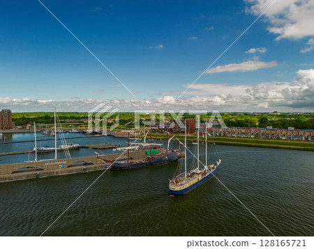 A large sailing ship cruises through the calm waters of a harbor under a clear blue sky. Other boats and yachts are docked nearby, with a town and green landscape visible in the background. 128165721