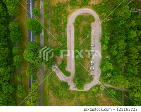 Aerial view of a small, curved forest parking area with several parked cars, surrounded by lush green trees and meadows, with a road running through the upper part of the image. 128165722