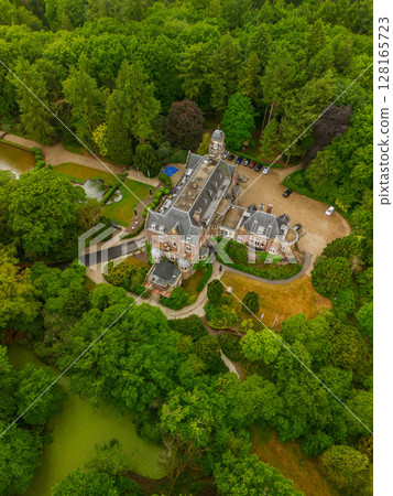 An aerial view of a historic mansion surrounded by lush green trees and a beautifully landscaped garden with a fountain, walking paths, and parking area. The scene shows vibrant summer greenery. 128165723