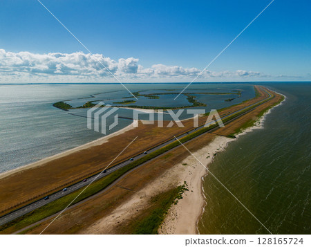Aerial view of a narrow dam or causeway flanked by expansive water bodies on both sides, leading toward the horizon under a vibrant blue sky with scattered clouds. 128165724