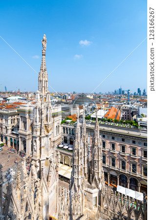 The stunning view from the rooftop of Duomo in Milan showcases ornate spires in the foreground and the modern skyline of the city under a clear blue sky. The stunning view from the rooftop of Duomo in Milan showcases ornate spires in the foreground and the modern skyline of the city under a clear blue sky. 128166267